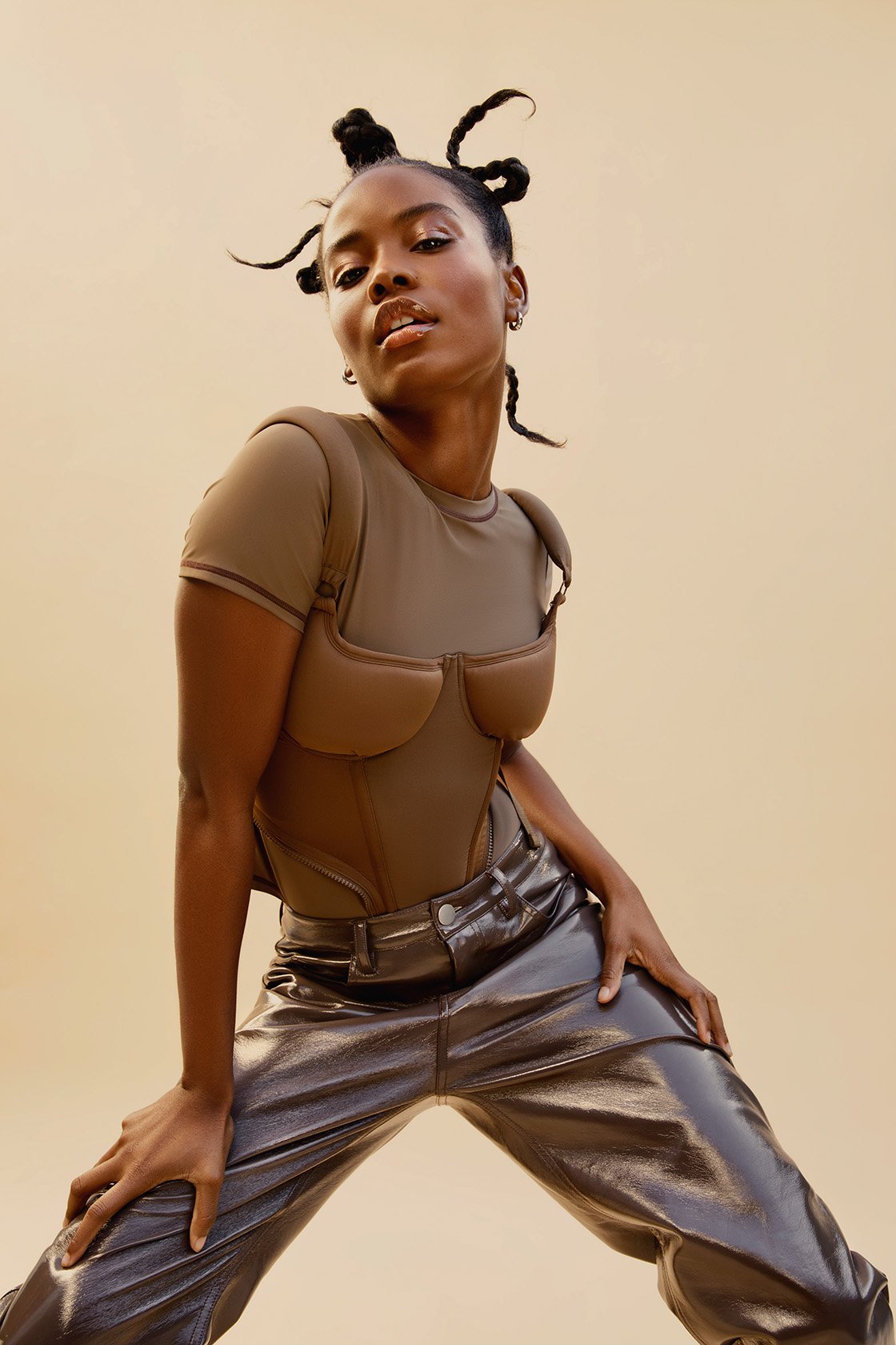 Fashionable woman in bodysuit and metallic pants poses in front of tan backdrop in studio. Dark-skinned model with twisted updo and hoop earrings.