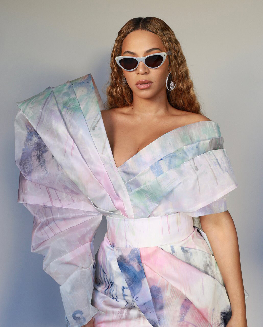 Beyonce wearing cat-eye sunglasses and an off-the-shoulder structured pastel dress against a plain grey background