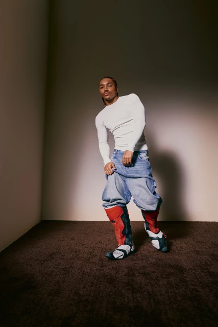An androgynous person poses in a photo studio wearing an off-white long-sleeve shirt, baggy light blue jeans, and red and black boots.