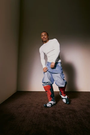 An androgynous person poses in a photo studio wearing an off-white long-sleeve shirt, baggy light blue jeans, and red and black boots.