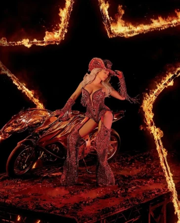 A woman in a red costume poses on a motorcycle with a flaming background. She wears a hat and an elaborate outfit.