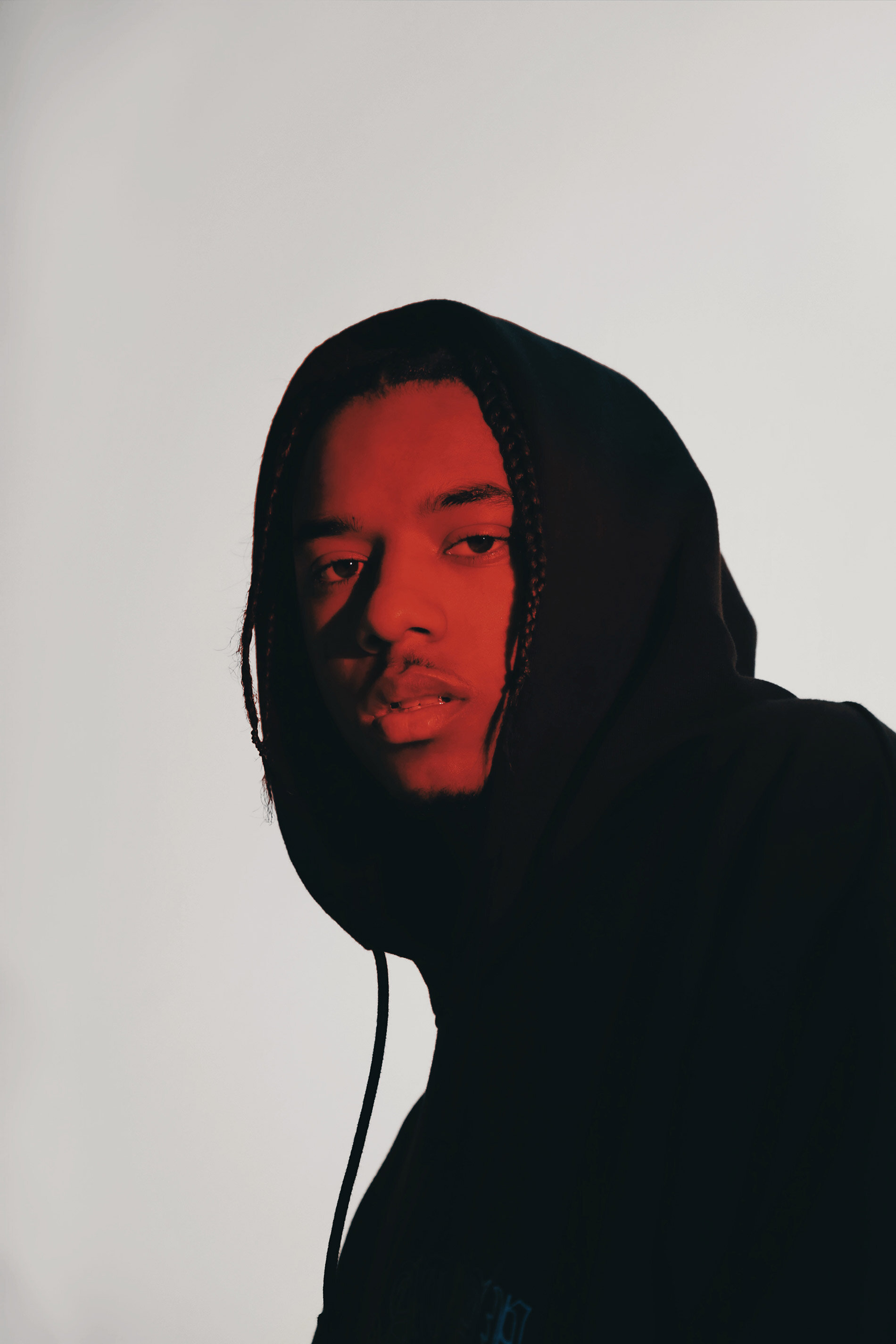 Portrait of an adult male with braided hair wearing a black hooded sweatshirt against a light grey backdrop with red lighting.