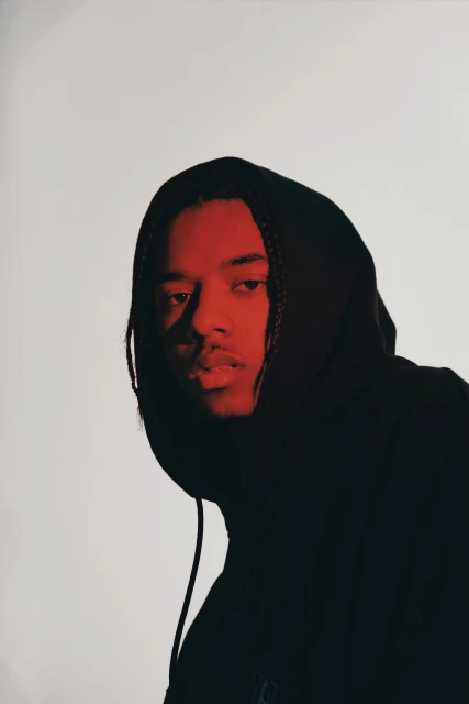 Portrait of an adult male with braided hair wearing a black hooded sweatshirt against a light grey backdrop with red lighting.
