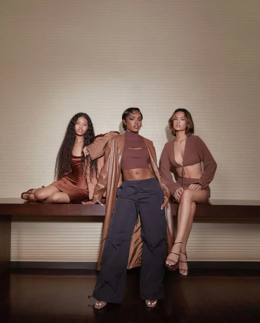 Three women pose indoors in stylish brown clothing. One wears a brown top and black wide-leg pants. The others wear brown dresses.