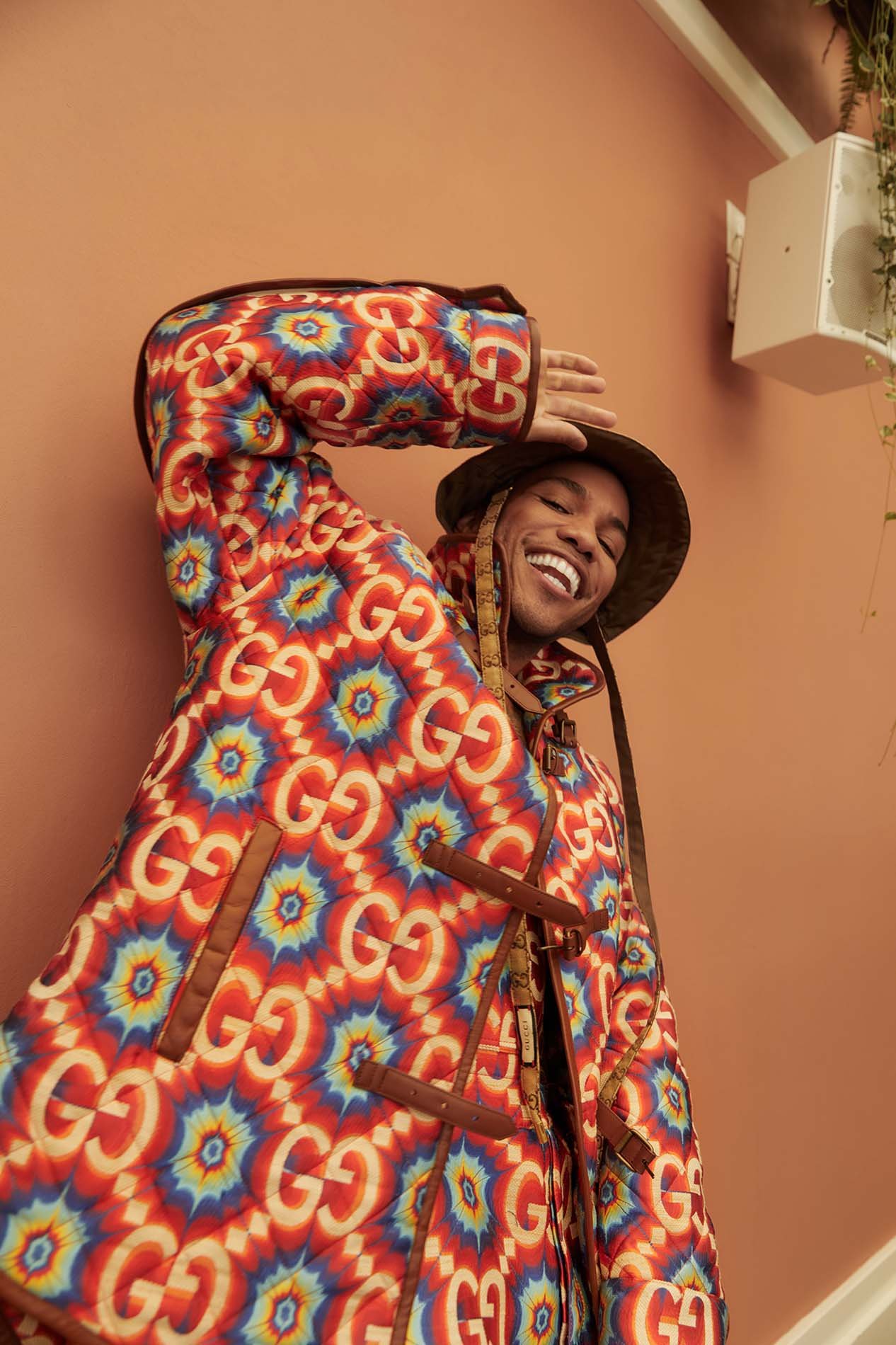Smiling adult in a patterned Gucci coat and hat poses indoors, with a light peach-colored wall in the background.