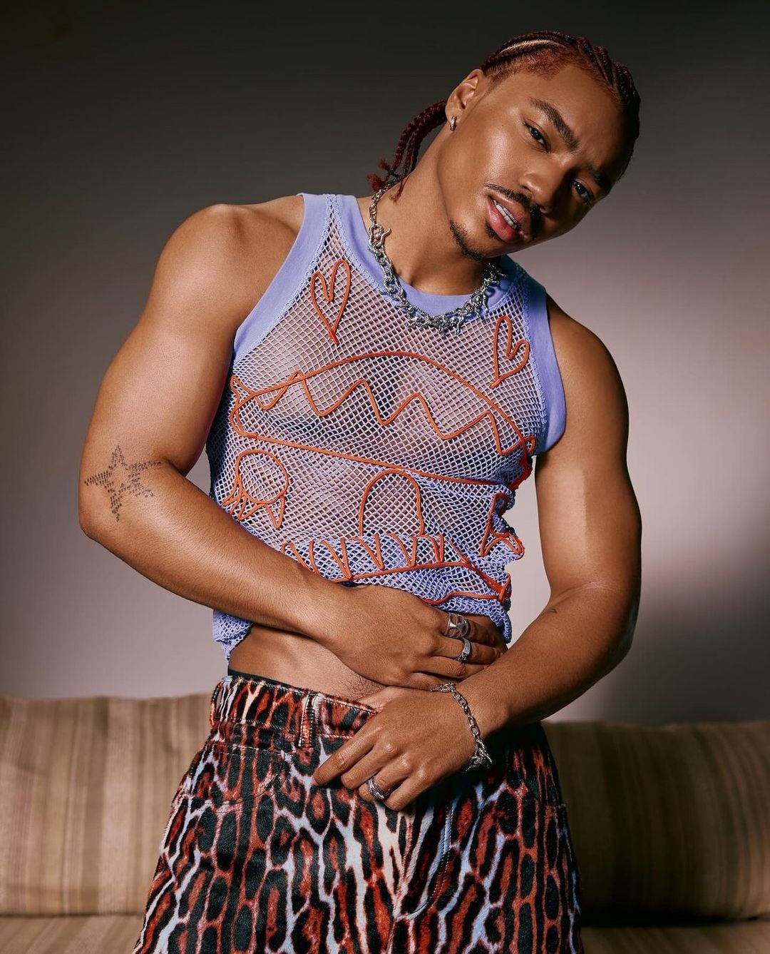 Portrait of a young adult male with braided hair, wearing a lilac-trimmed mesh tank top, animal print skirt, and jewelry against a gray backdrop.
