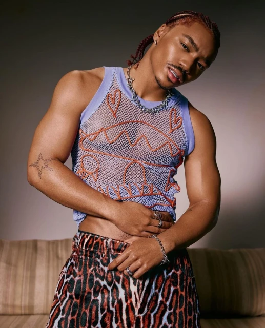 Portrait of a young adult male with braided hair, wearing a lilac-trimmed mesh tank top, animal print skirt, and jewelry against a gray backdrop.