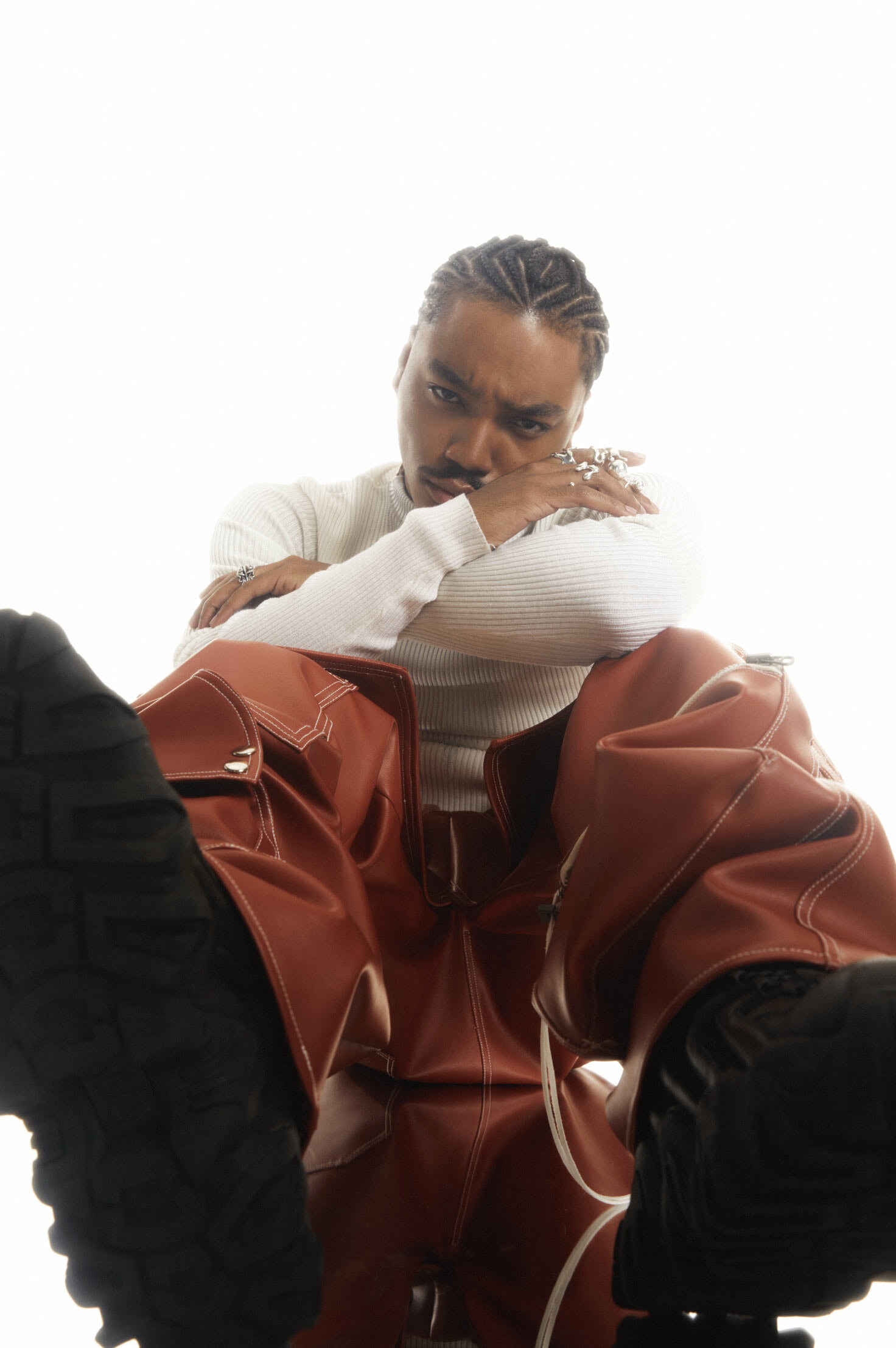 Portrait of a stylish adult male with braids wearing a white sweater and rust-colored pants posing against a white background.