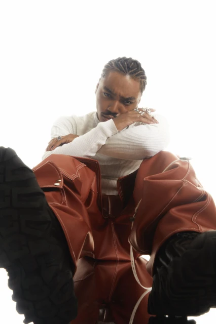 Portrait of a stylish adult male with braids wearing a white sweater and rust-colored pants posing against a white background.