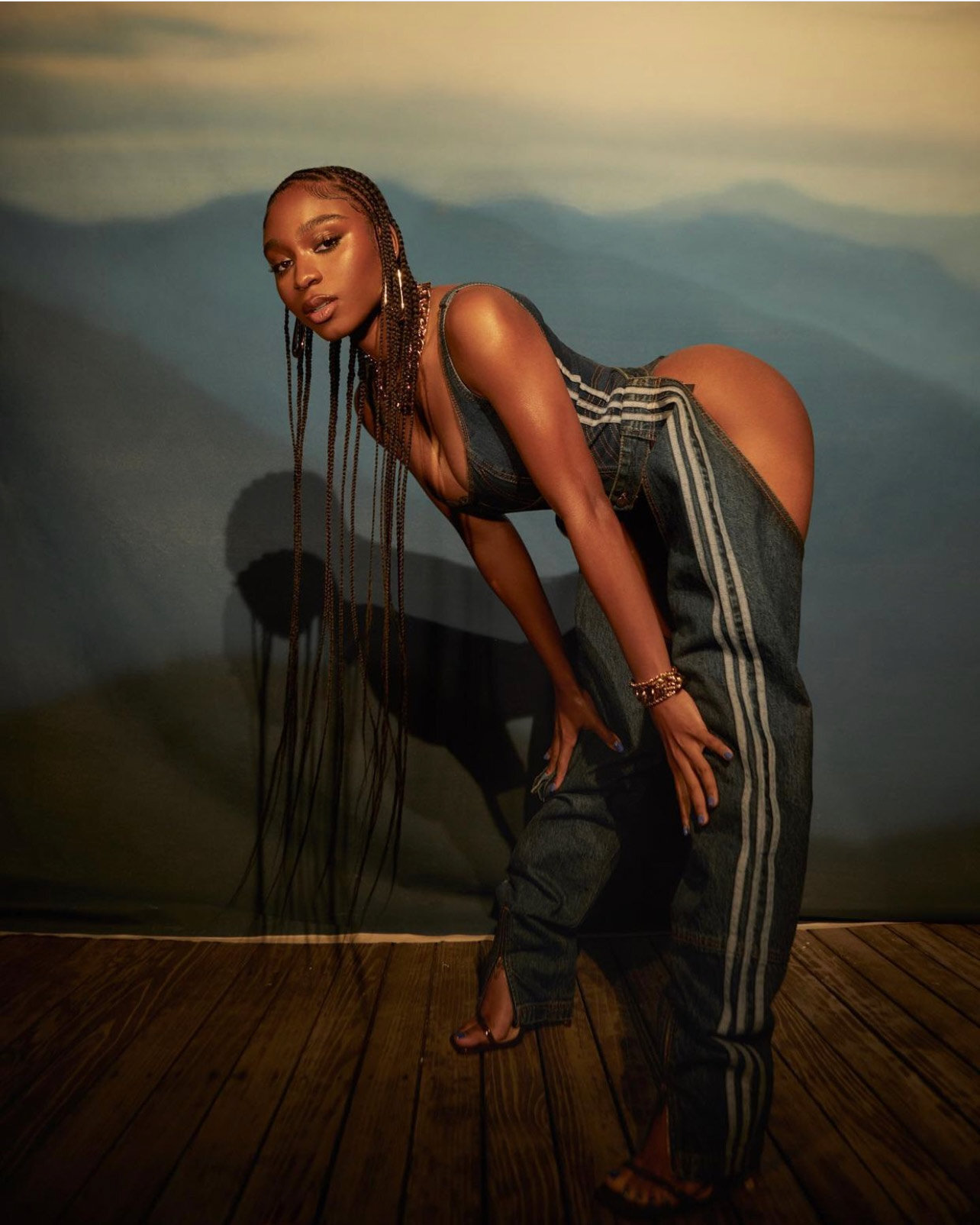 Normani with long braided hair wearing denim chaps and top posed against a painted mountain backdrop by Blair Caldwell