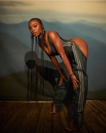 Normani with long braided hair wearing denim chaps and top posed against a painted mountain backdrop by Blair Caldwell