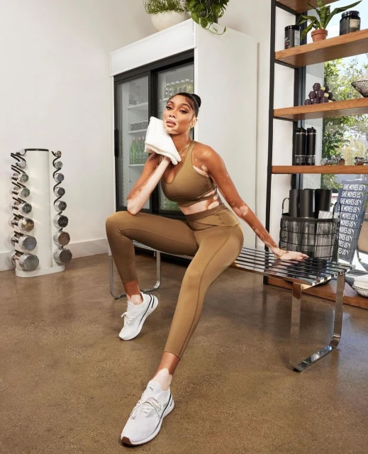 Athletic woman with vitiligo sits in workout gear, holding towel, in a gym setting with weights and water bottles visible.
