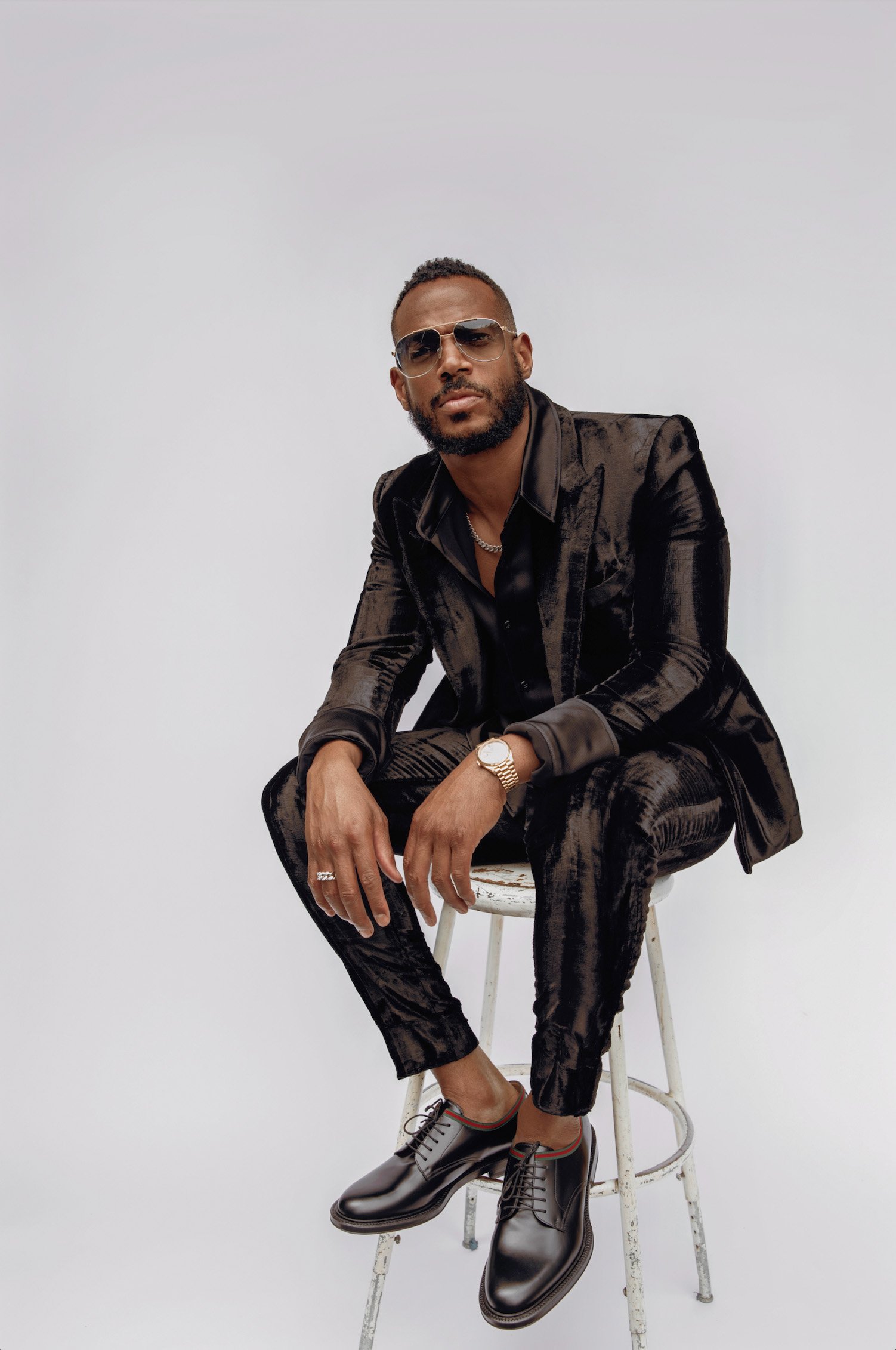 Portrait of a dark-skinned man in a crushed velvet suit sitting on a stool, wearing sunglasses, watch, and dress shoes, against a white backdrop.