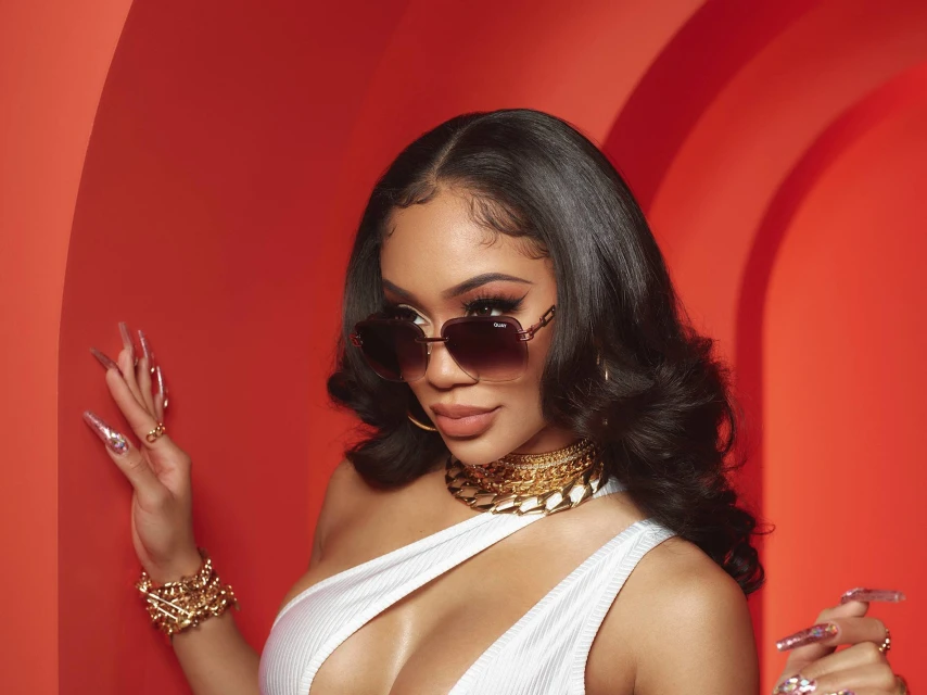 Portrait of an adult female with long dark hair, wearing sunglasses, jewelry, and a white top against a red background.