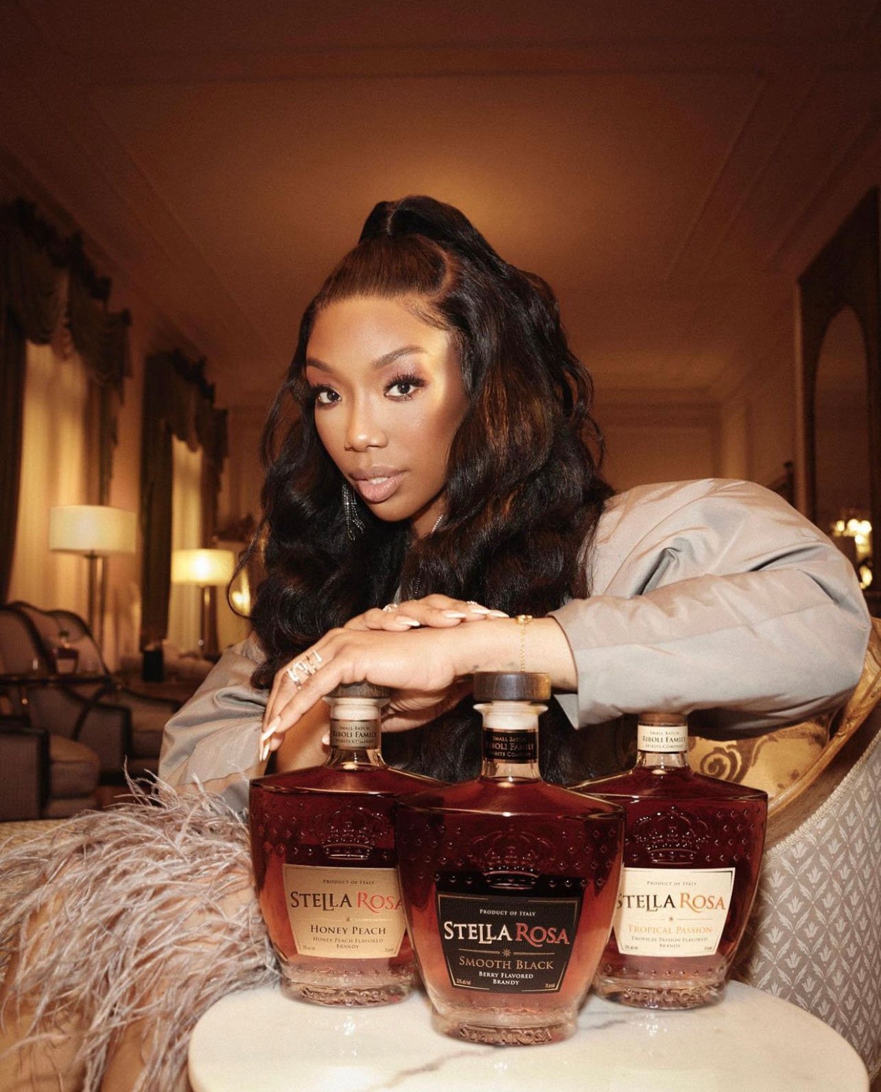 Brandy Norwood poses with Stella Rosa beverages in a warm-toned setting, wearing a grey blazer and diamond jewelry. Soft, elegant atmosphere.
