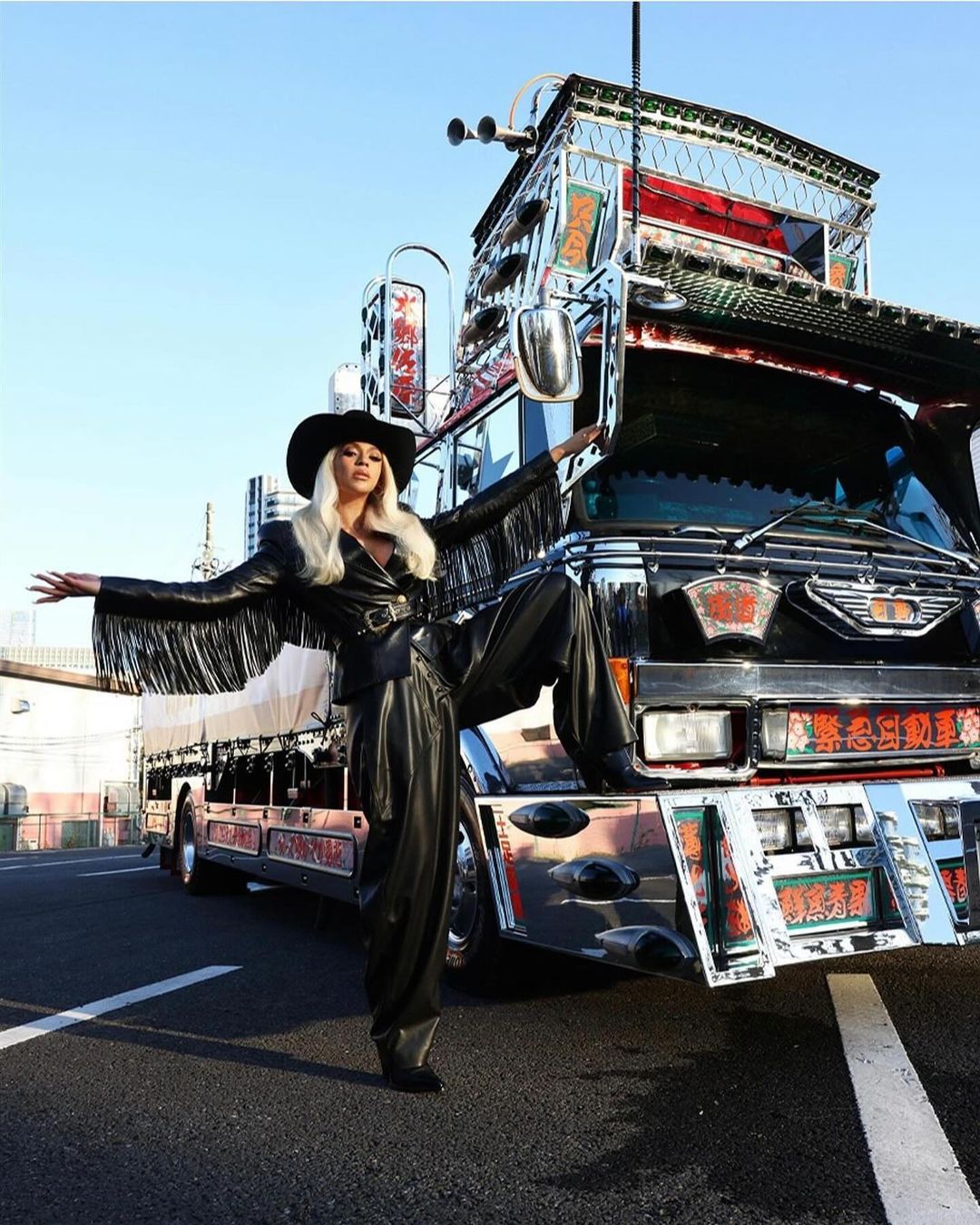 Stylish person in black hat and fringed jacket poses on customized truck, creating a bold fashion statement against urban background.