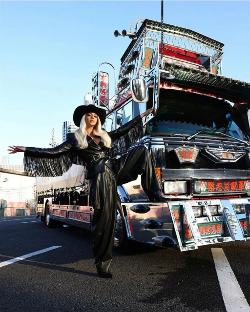 Stylish person in black hat and fringed jacket poses on customized truck, creating a bold fashion statement against urban background.