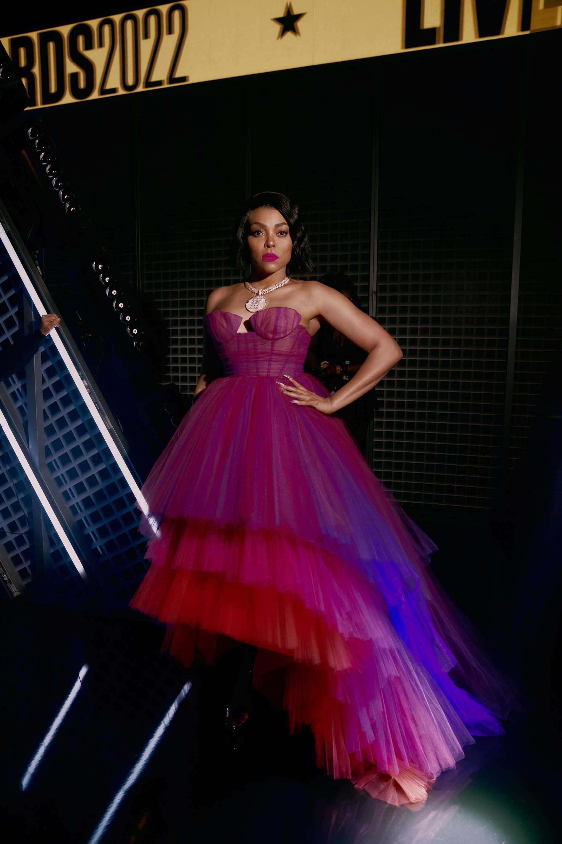 Taraji P. Henson poses on a red carpet in a pink and red dress with a tiered skirt at the BET Awards 2022, smiling, medium shot.