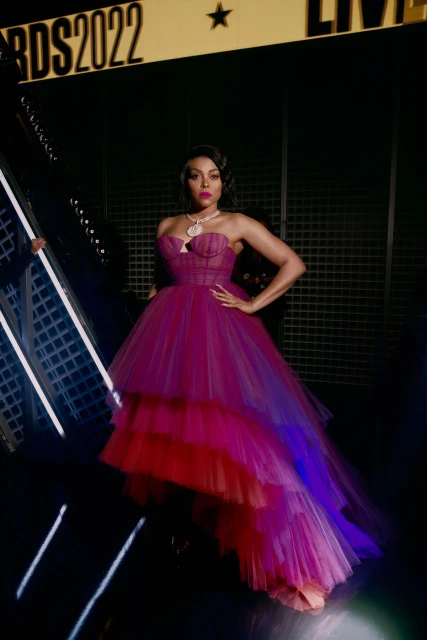 Taraji P. Henson poses on a red carpet in a pink and red dress with a tiered skirt at the BET Awards 2022, smiling, medium shot.