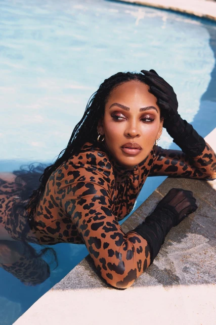 Adult female rests in a pool wearing an animal-print bodysuit and sheer gloves. She has long, dark hair, bronze makeup, and gold hoops.