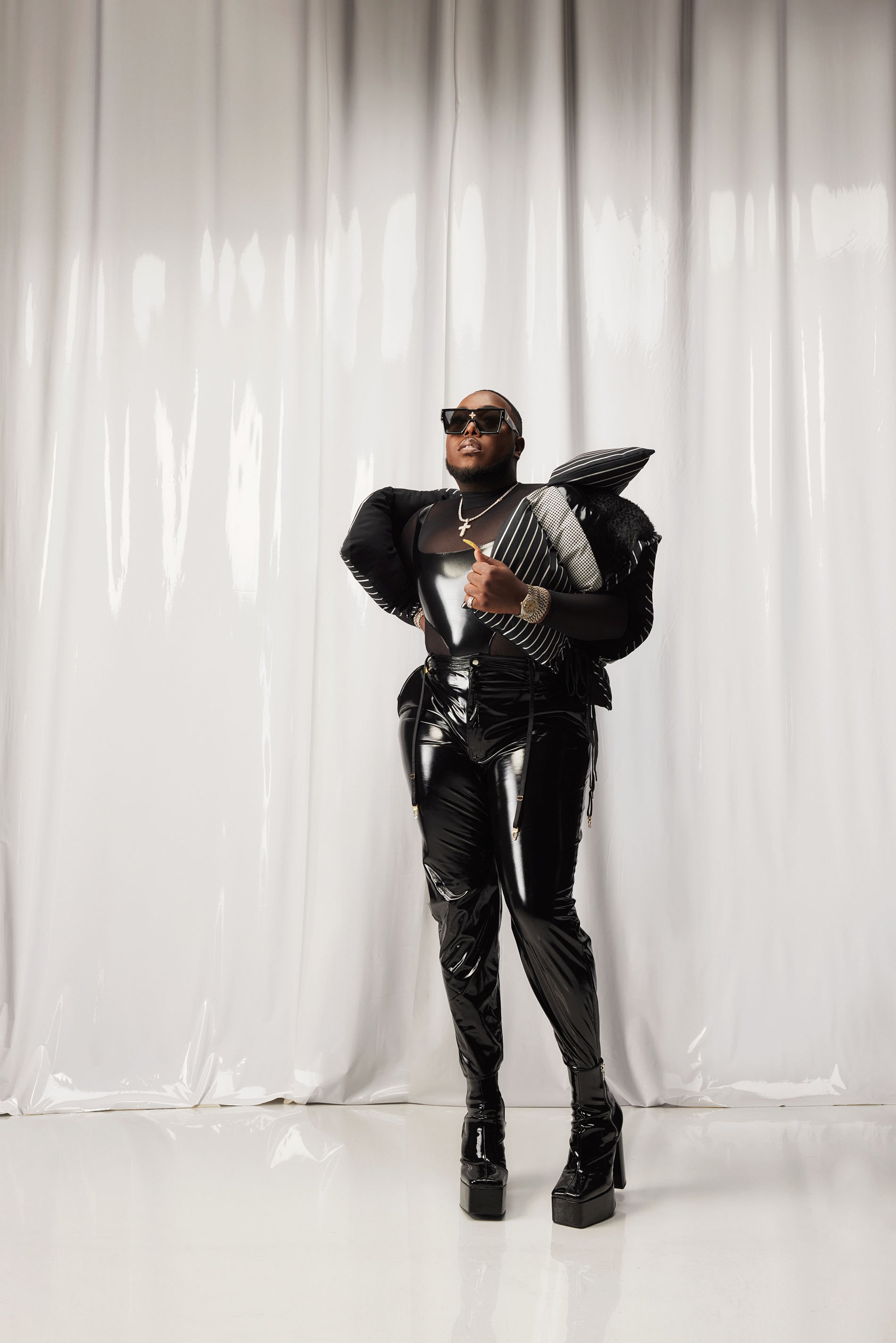 Adult male in a black shiny outfit and platform boots posing in a photo studio with a white curtain backdrop and studio lighting.