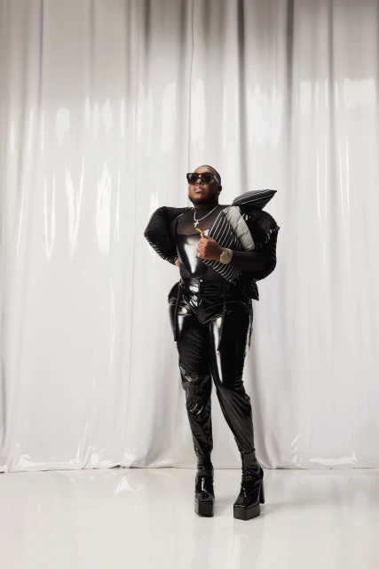 Adult male in a black shiny outfit and platform boots posing in a photo studio with a white curtain backdrop and studio lighting.