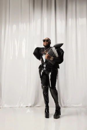 Adult male in a black shiny outfit and platform boots posing in a photo studio with a white curtain backdrop and studio lighting.