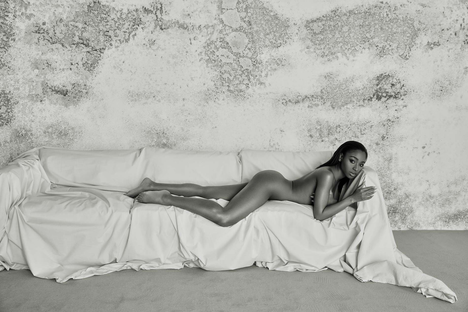 Monochrome studio shot of an adult woman lying on a white couch in a reclined pose, with a textured wall visible in the background.