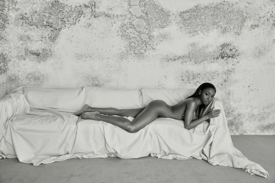 Monochrome studio shot of an adult woman lying on a white couch in a reclined pose, with a textured wall visible in the background.