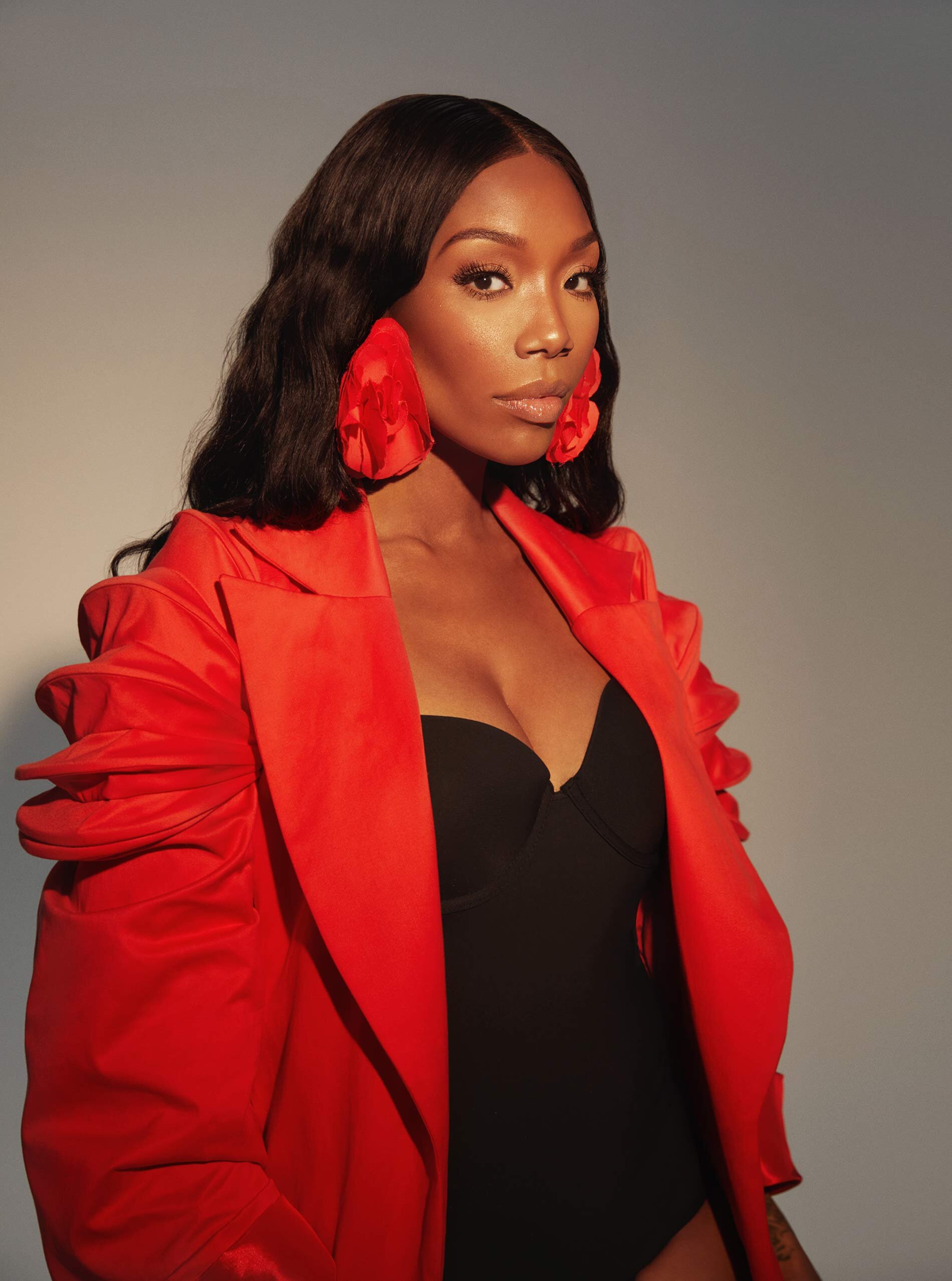 Portrait of a woman with long, dark hair wearing a black top, a bright red oversized jacket, and large red flower-shaped earrings.