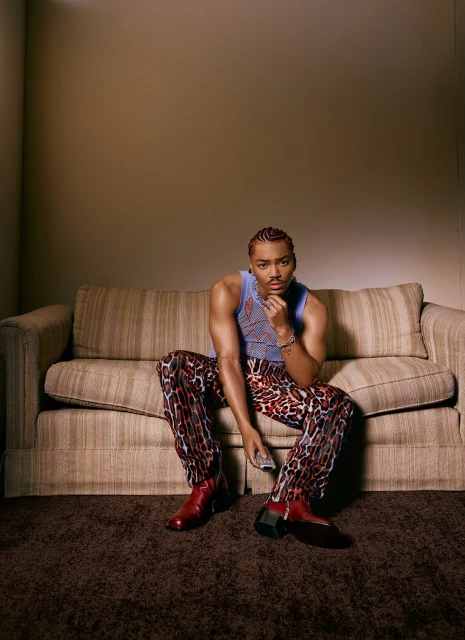 A young Black man sits on a sofa in a photo studio wearing a blue tank top, leopard-print pants, and red boots, holding a remote control.