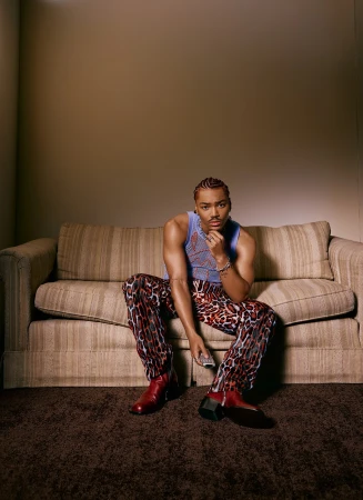 A young Black man sits on a sofa in a photo studio wearing a blue tank top, leopard-print pants, and red boots, holding a remote control.