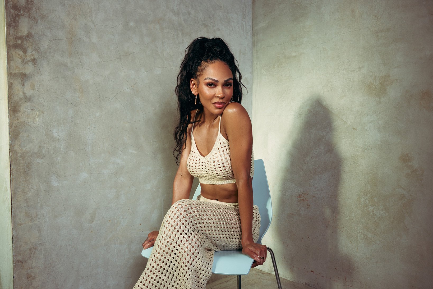 Attractive Black woman with long hair poses in a knit crop top and pants while sitting on a chair against a textured wall in studio light.