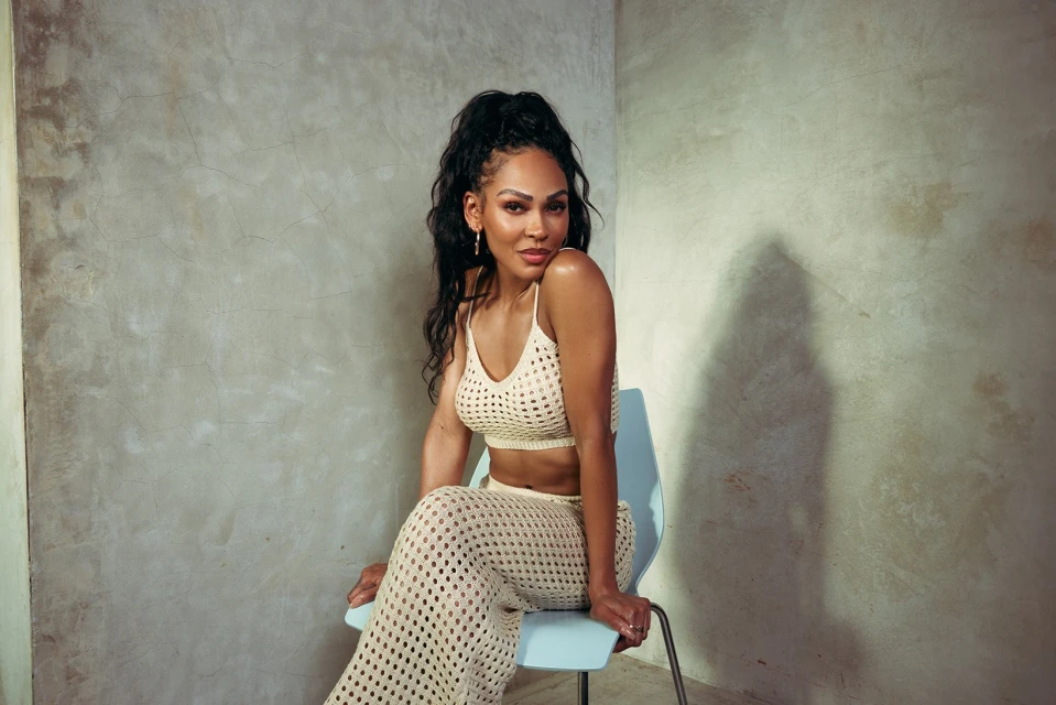 Attractive Black woman with long hair poses in a knit crop top and pants while sitting on a chair against a textured wall in studio light.