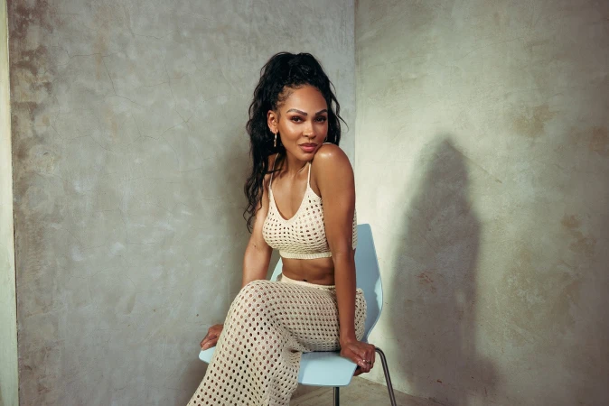 Attractive Black woman with long hair poses in a knit crop top and pants while sitting on a chair against a textured wall in studio light.