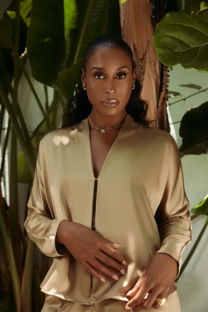 Portrait of a stylish adult woman with dark hair posing in front of green foliage. She is wearing a tan colored satin shirt and pants.