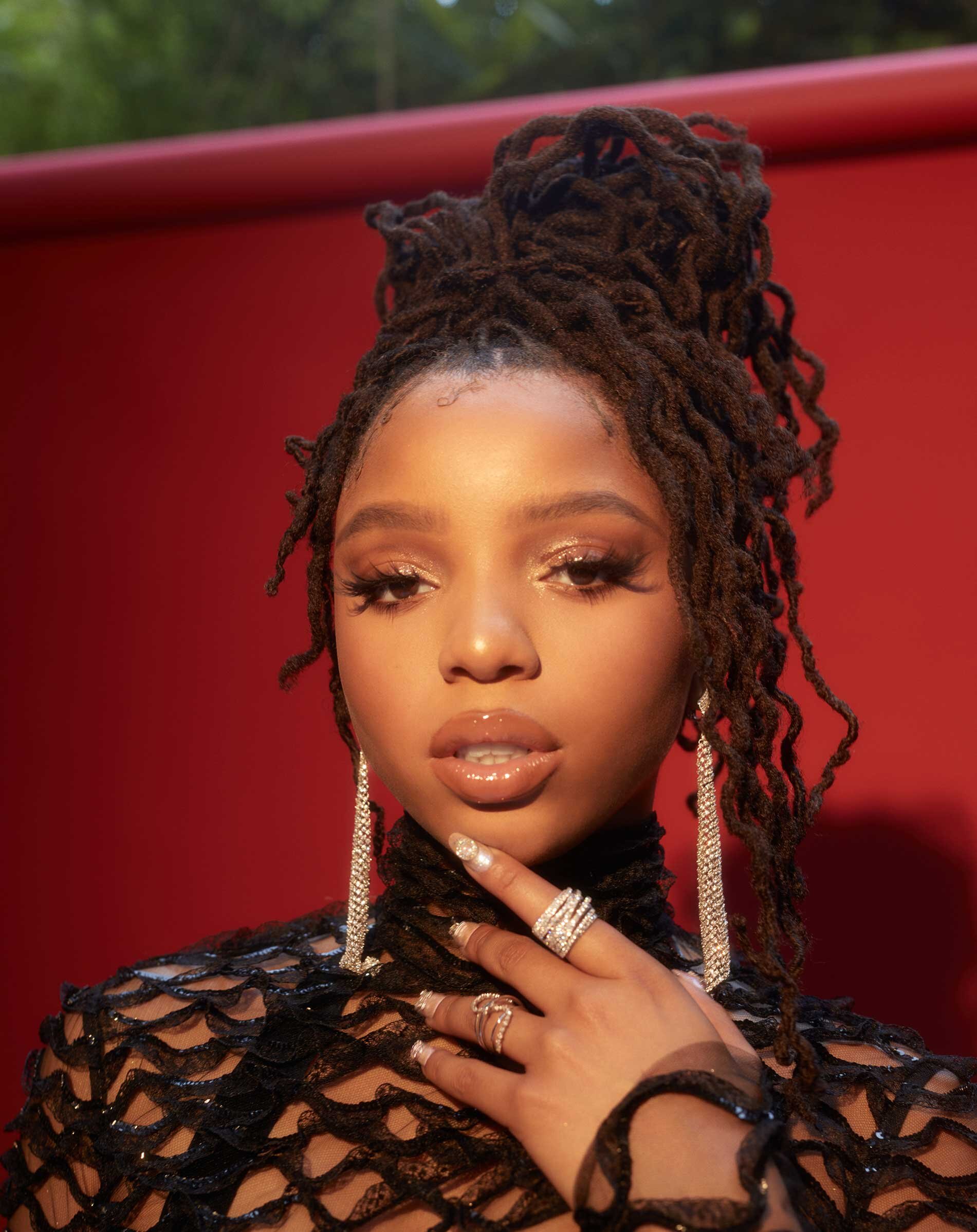 Close-up portrait of an adult female with brown locs styled in a high bun, posing against a red background, wearing a black mesh top.