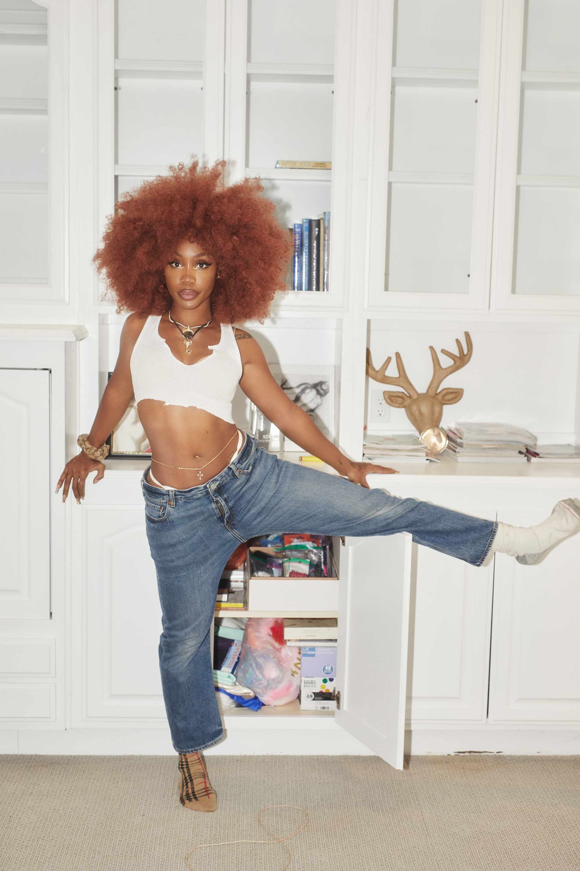Woman with large afro, wearing a white crop top and jeans, poses with one leg inside a white cupboard in an indoor setting.