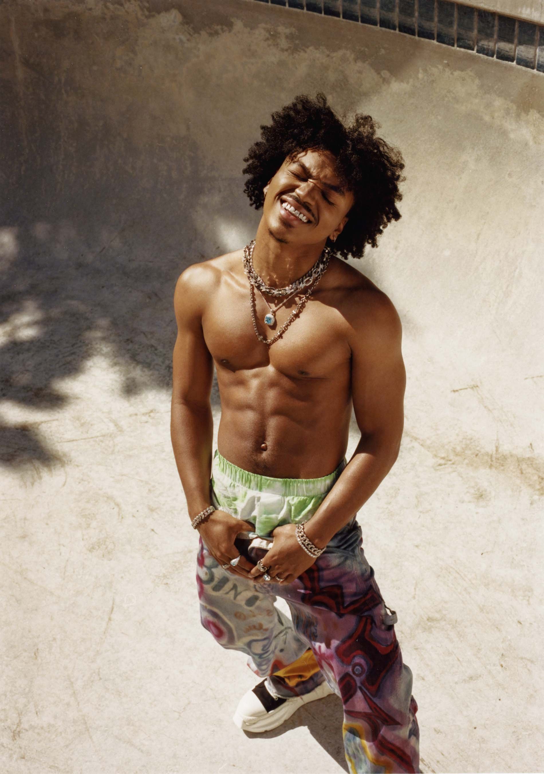 Shirtless man with curly hair smiles, wearing jewelry and patterned pants, stands in a concrete bowl with sunlight, showing his physique.