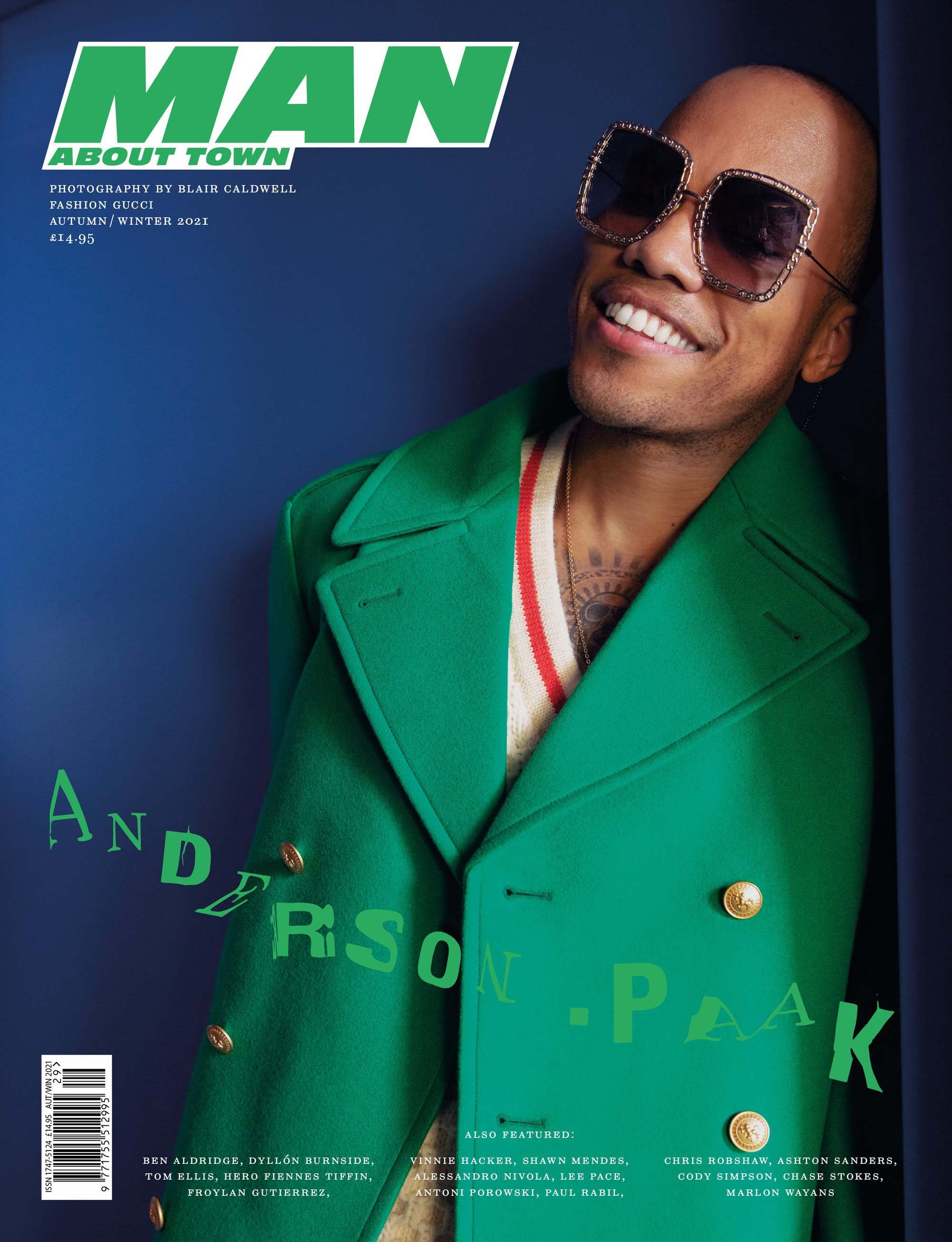 Magazine cover featuring a smiling adult male wearing large sunglasses and a green coat against a dark blue background with text.