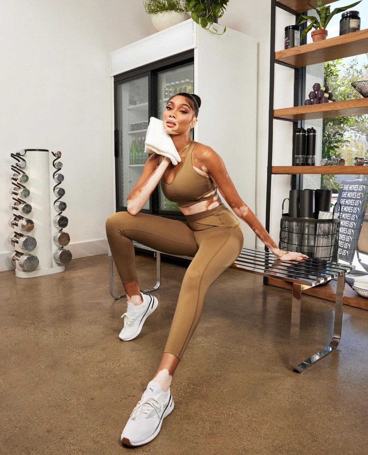 An adult female model in athletic wear sits on a metal bench in a gym setting, holding a towel, showcasing a modern fitness aesthetic.
