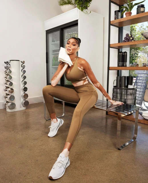 An adult female model in athletic wear sits on a metal bench in a gym setting, holding a towel, showcasing a modern fitness aesthetic.