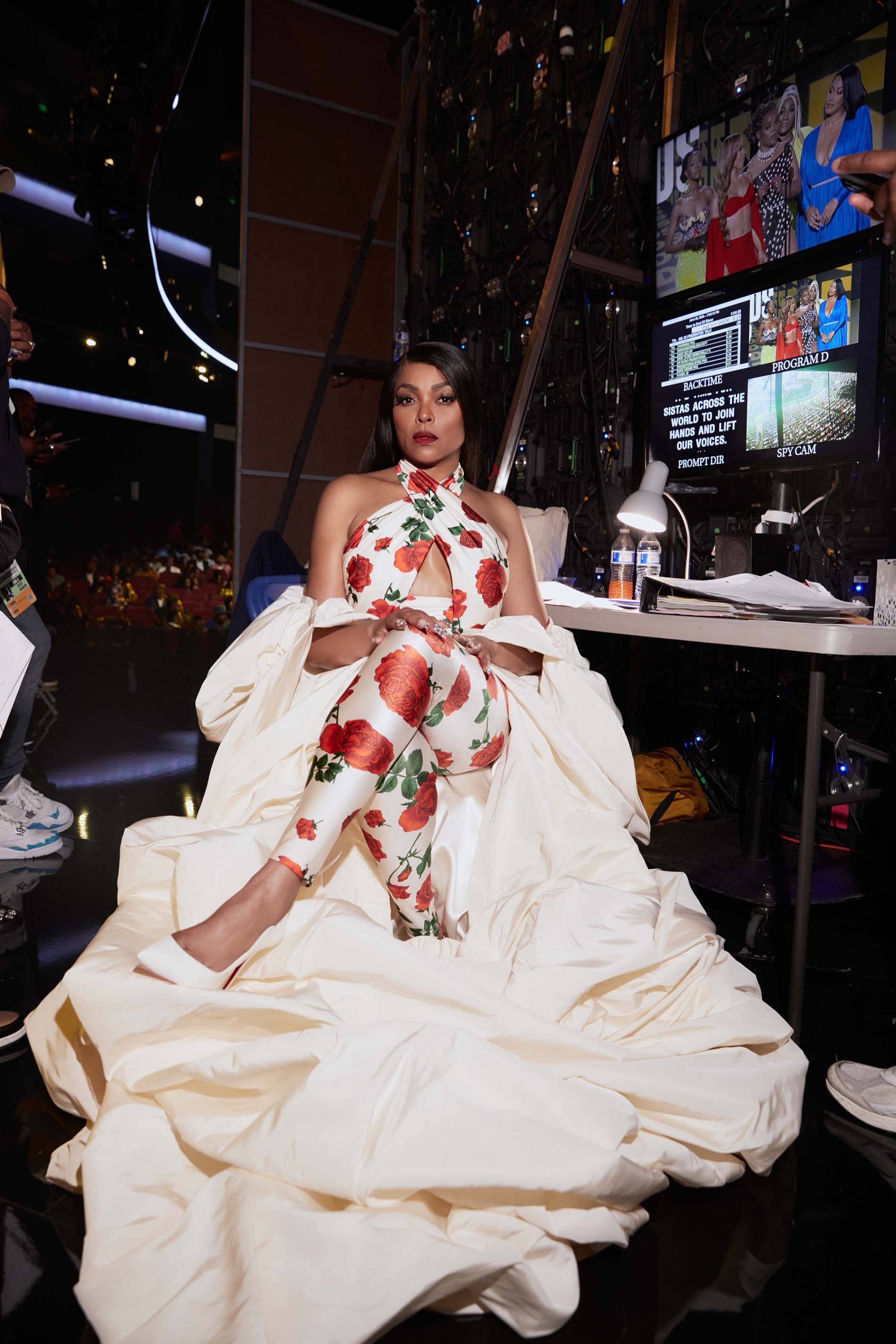 Taraji P. Henson sits wearing a floral outfit with a cream-colored gown, at a desk with monitors in a backstage setting.
