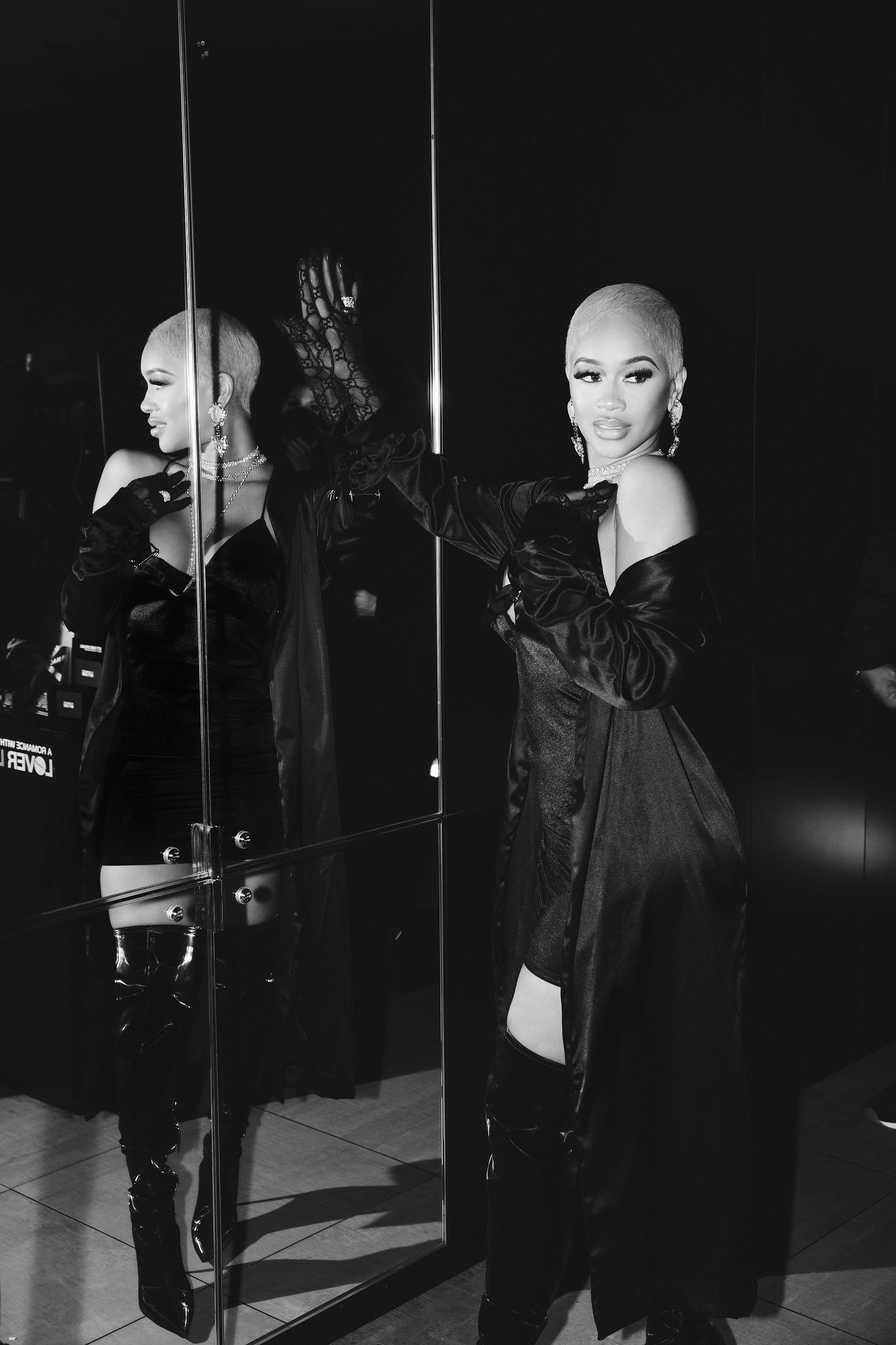 Black-and-white shot of a bald female posing by a mirror, wearing thigh-high boots, a dark dress and gloves in a dark studio.