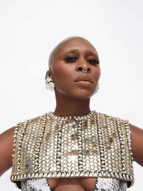 Close-up studio shot of a bald adult woman with a nose ring, wearing a jeweled top with dramatic earrings against a bright white background.