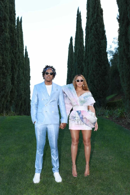 Beyonce and Jay-Z posing together on a grassy lawn with tall, slender trees in the background, both wearing fashionable outfits. 