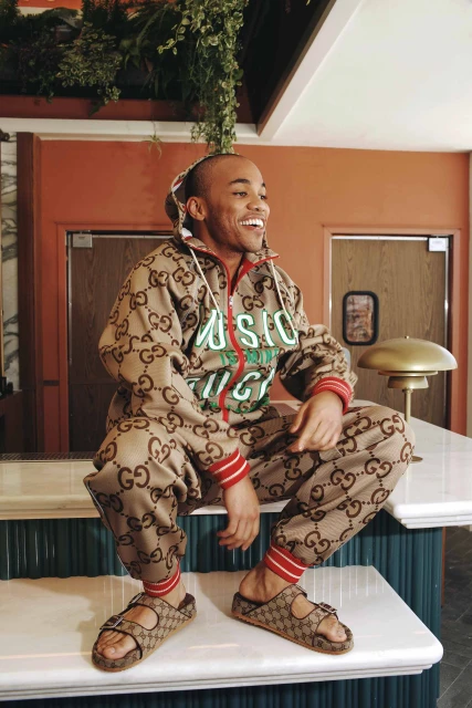Smiling adult man in a patterned Gucci tracksuit and sandals sitting on a white table with foliage in the background.