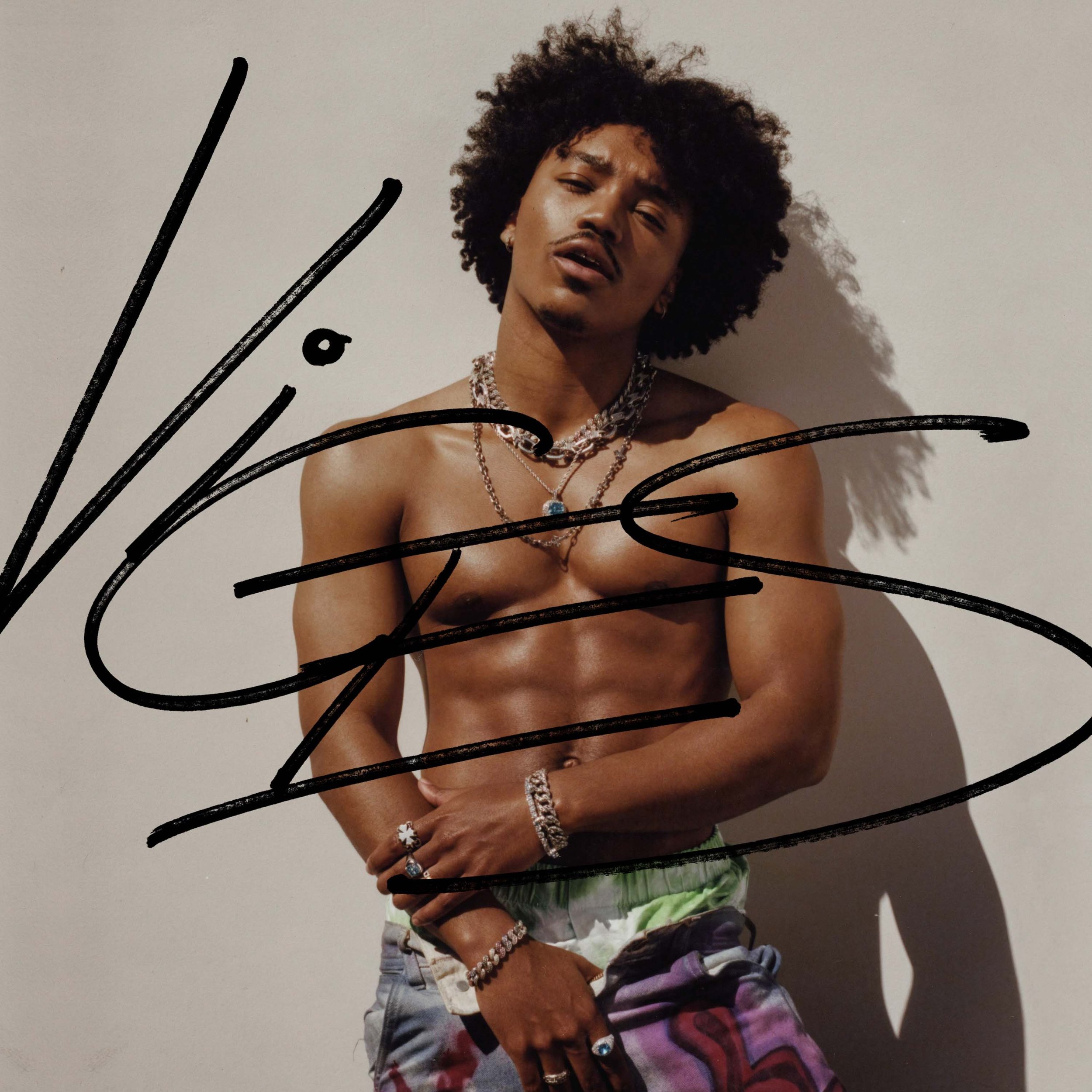 Shirtless man with curly hair, wearing bracelets and necklaces, stands indoors against a plain wall in a medium shot.