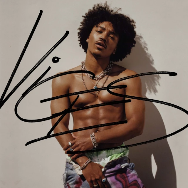 Shirtless man with curly hair, wearing bracelets and necklaces, stands indoors against a plain wall in a medium shot.