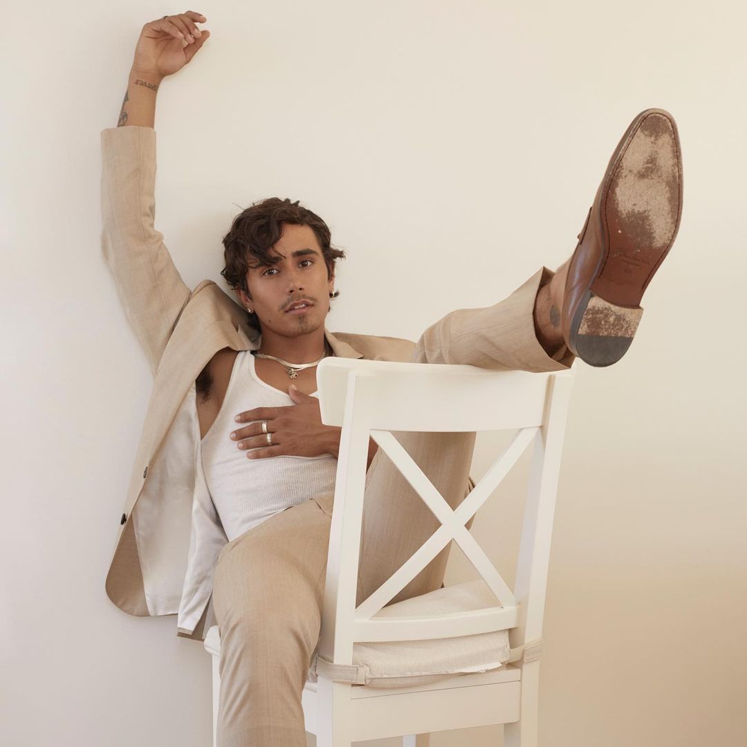 Young adult man posing casually on a chair in a beige suit, white tank top, and dress shoes in an indoor setting against a neutral wall.